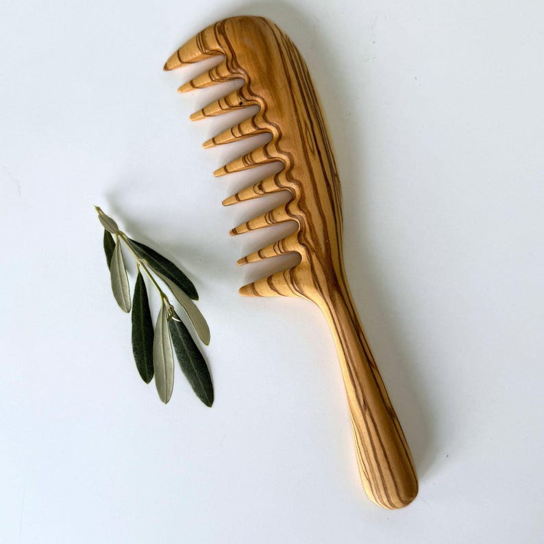May include: A wooden comb with a natural wood grain pattern. The comb has a wide handle and a row of teeth. The comb is made of olive wood.