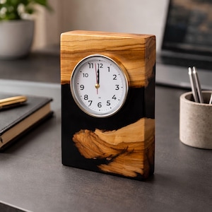 May include: A rectangular desk clock featuring a natural light brown wood grain body contrasted with a smooth black resin section. The clock face is white with black Arabic numerals, a silver-toned circular frame, and three thin hands.