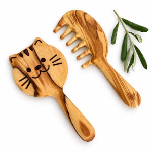 Pode incluir: Uma escova de cabelo de madeira castanho-claro com uma gravação de cara de gato e um pente de madeira a condizer, ambos com um padrão de grão de madeira natural, acompanhados por um pequeno ramo de oliveira verde com folhas.