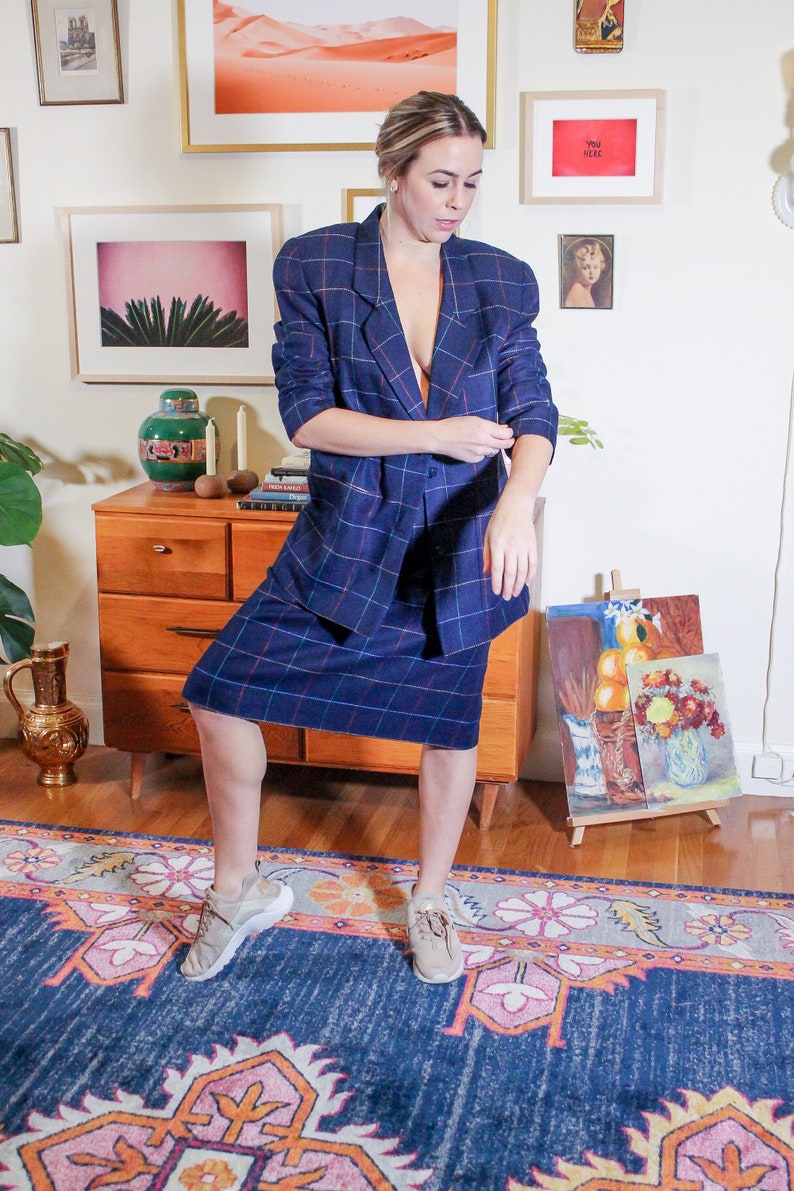 May include: A woman wearing a blue plaid blazer and skirt, standing on a blue and pink patterned rug. She is wearing beige sneakers and has her hair pulled back.