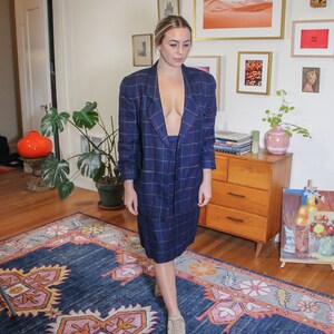 May include: A woman wearing a blue and white plaid blazer and skirt set. She is standing on a blue and pink patterned rug in a room with a dresser and artwork on the walls.