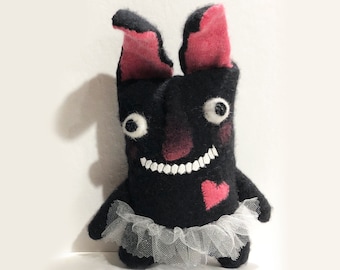 May include: Black felt bunny with pink ears, a white tulle tutu, and a pink heart on its chest. The bunny has white eyes and a wide, toothy smile.