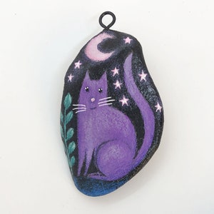 May include: Purple cat painted on a rock with a crescent moon and stars. The rock has a black wire loop for hanging.