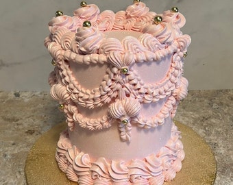 Puede incluir: Un pastel rosa decorado con cuentas doradas y tuberías intrincadas. El pastel está sobre una base de pastel de brillo dorado.