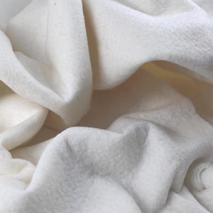 May include: Close-up of a soft, off-white fabric with a textured surface. The fabric is draped and folded, creating shadows and highlights. The material appears to be thick and possibly used for clothing or home decor.