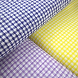 May include: A close-up shot of three rolls of gingham fabric in blue, yellow, and purple. The fabric has a classic checked pattern, ideal for sewing. The colours are bright and the material appears to be cotton, suitable for various craft projects.