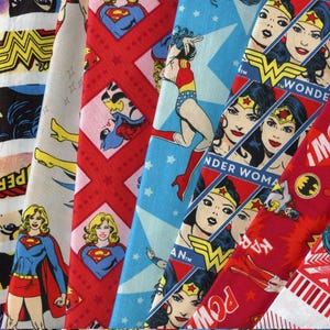 May include: A selection of patterned fabrics featuring comic book superheroes. Designs include Wonder Woman, Superman, and Batman. The fabrics display various patterns, including stars, diamonds, and character portraits. The colour palette includes red, blue, yellow, and white.