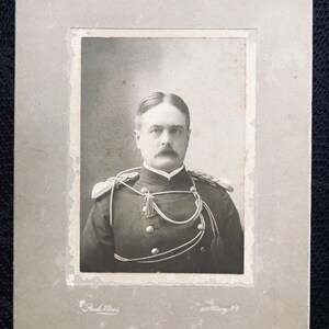 May include: Black and white portrait photograph of a man in a military uniform. He is wearing a dark jacket with gold buttons and a white shirt. He has a mustache and a serious expression. The photo is dated "1897".