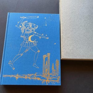 May include: A blue book with gold illustrations of a mythological figure and celestial elements. The book is in a matching gray slipcase. The cover art features a figure with a ram's head, a crescent moon, and stars.