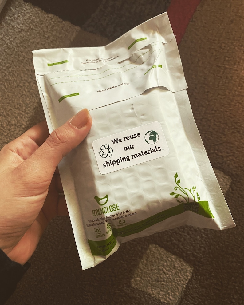 Może przedstawiać: Biała koperta wysyłkowa z zielono-białą naklejką z napisem "Ponownie wykorzystujemy nasze materiały wysyłkowe". Koperta jest wykonana z recyklingowanego papieru bąbelkowego i ma wymiary 10,16 cm x 17,15 cm.