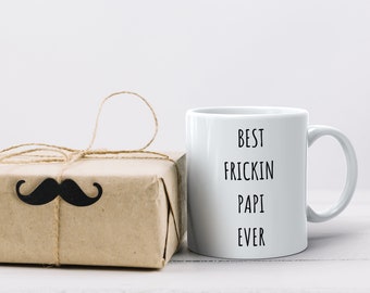 May include: White ceramic mug with a black handwritten message that reads "Best Frickin Papi Ever". A brown gift box with a black mustache is in the background.