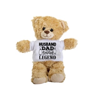 May include: A brown teddy bear wearing a white t-shirt with the text "Husband Dad Bowling Legend" and two black bowling pins on either side of the text.