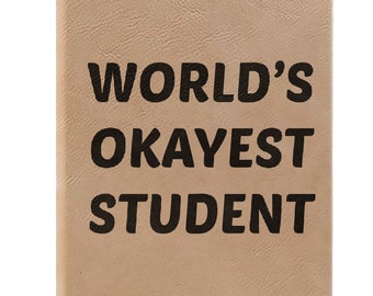 May include: A tan leather-bound notebook with the text "WORLD'S OKAYEST STUDENT" printed in black.
