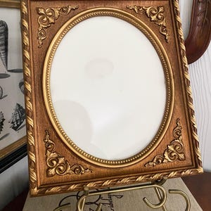 May include: A rectangular picture frame with a gold finish and an oval center opening. The frame features ornate corner scrollwork, a beaded inner border, and a twisted rope outer edge. It is displayed on a gold-toned metal wire stand.