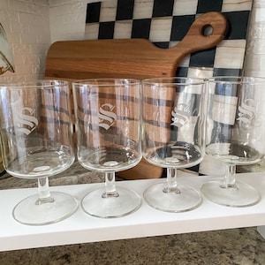 May include: Four clear glass goblets with a stem and a round base. Each glass has a large, stylized letter "S" etched on the side. The glasses are displayed on a white shelf.