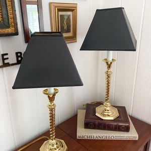 May include: Two brass table lamps with black square shades. The lamps have a twisted column design and sit on a dark wooden table. The lamps are next to a stack of books, including one titled "MICHELANGELO".