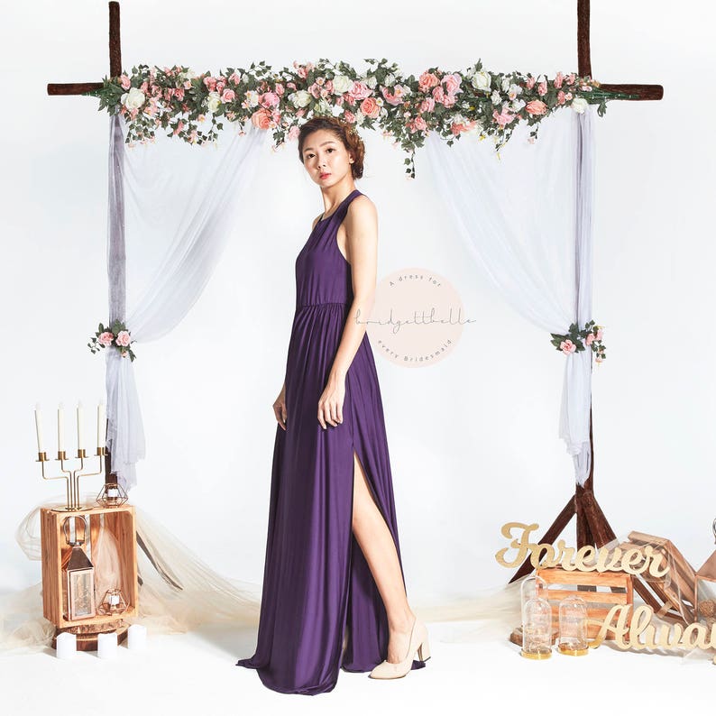 May include: A woman wearing a long, purple maxi dress with a side slit. The dress has a halter neckline and a fitted bodice. She is standing in front of a floral archway with a white sheer curtain behind it. The text "A Girl For Every Bride" and "Ladygathalle" is visible on the image.