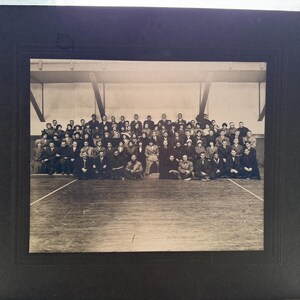 May include: A black and white photograph of a large group of people, likely a class or team, posing indoors. The photo is in a black frame, with the group arranged on a wooden floor in front of a wall.
