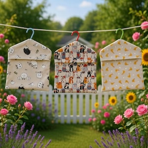 May include: Three patterned fabric pouches hang from a washing line. One features sheep, another cats, and the third has yellow shapes. Each pouch is suspended by a colourful plastic hanger, set against a backdrop of flowers and greenery.