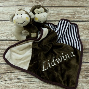 Op de afbeelding: Een bruine en beige babydeken met een zwart-wit gestreepte rand. Twee bruine aapjes van pluche zijn aan de deken bevestigd. De deken is gepersonaliseerd met de naam "Lidwina" geborduurd in wit.