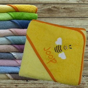 Könnte beinhalten: Ein Stapel gefalteter Kapuzenhandtücher in verschiedenen Farben wie Gelb, Grün, Blau, Rosa und Braun. Das oberste gelbe Handtuch zeigt eine gestickte Biene und das Wort Joop in orangefarbenem Garn mit einem passenden orangefarbenen Rand.