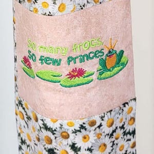 Könnte beinhalten: Eine Stofftasche mit Gänseblümchenmuster und einem rosa Panel mit einer gestickten Fröschen und dem Text "So many frogs, so few princes." Die Tasche hat einen gelben Riemen.