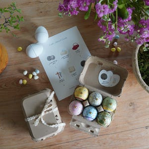 May include: An assortment of Easter-themed items on a wooden surface. Includes a carton of decorated eggs, a closed egg carton tied with lace, white eggs, a card with illustrations, and purple flowers in a pot. Small, colorful candies are scattered around.