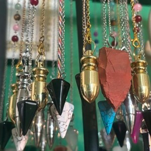 May include: A collection of hanging pendulums featuring various materials including polished gold and silver metal caps, smooth black stone points, white marble-like stone points, a rough red stone point, and a bright blue turquoise-colored stone point.