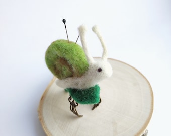 Puede incluir: Un caracol de fieltro blanco con una concha de fieltro verde y patas de bronce. El caracol está sentado sobre una superficie de madera.