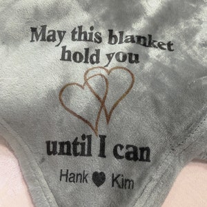 Puede incluir: Una manta de franela gris con el texto "May this blanket hold you until I can Hank ♥ Kim" impreso en ella. Dos corazones dorados están impresos sobre el texto.