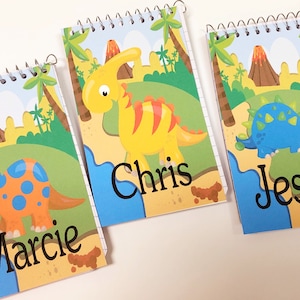 May include: Three spiral-bound notebooks with dinosaur illustrations. Each notebook features a different dinosaur in vibrant colors against a prehistoric landscape. The names "Marcie", "Chris", and "Jessie" are printed on the covers.