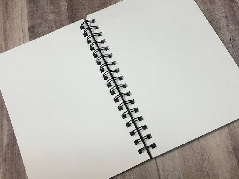 Op de afbeelding: Een open, blanco spiraalgebonden notitieboek met witte pagina's. De zwarte metalen spiraalbinding is zichtbaar aan de linkerkant. Het notitieboek staat schuin op een lichtbruin houten oppervlak.