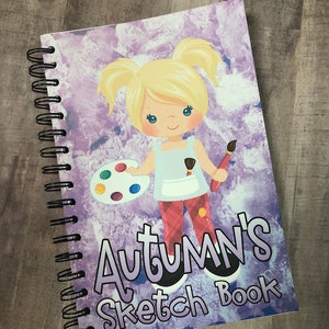 Op de afbeelding: Spiraalgebonden schetsboek met een omslag met een cartoonmeisje met een penseel en palet. De omslag heeft de tekst "Autumn's Sketch Book" in een paars en wit ontwerp. De achtergrond is een paars en wit aquareleffect.