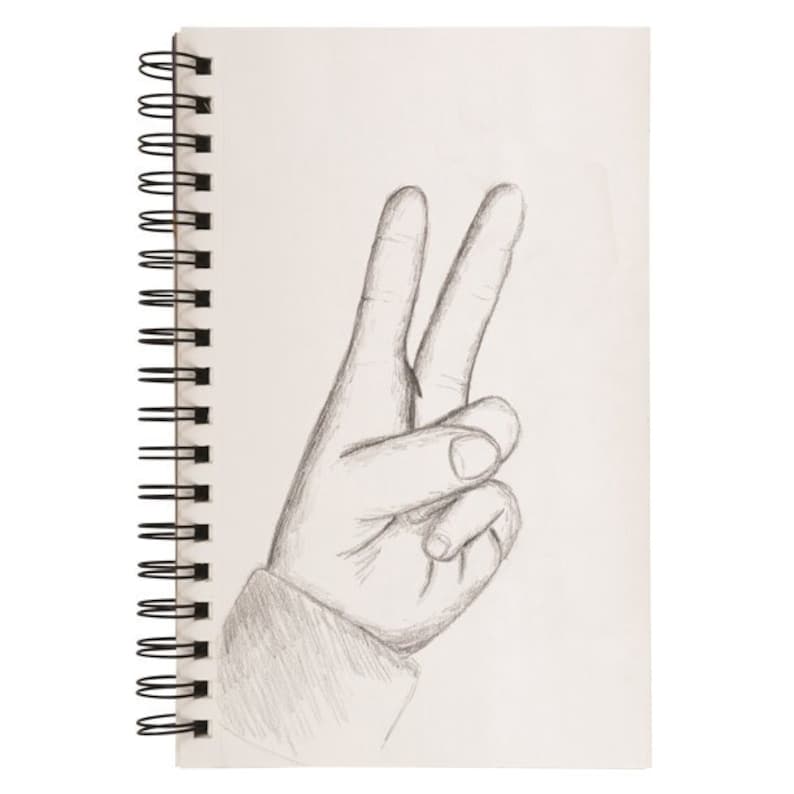 Op de afbeelding: Een spiraalgebonden schetsboek met een potloodschets van een hand die een vredesteken maakt. De tekening is in grijstinten, met de vingers uitgestrekt om de V-vorm te vormen. De pagina's van het schetsboek zijn wit.