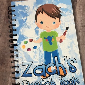 Op de afbeelding: Een spiraalgebonden schetsboek met een blauw en wit hemelthema. De omslag toont een cartoonillustratie van een jongen met een penseel en een verfpalet. De tekst "Zach's Sketch Book" staat op de omslag.