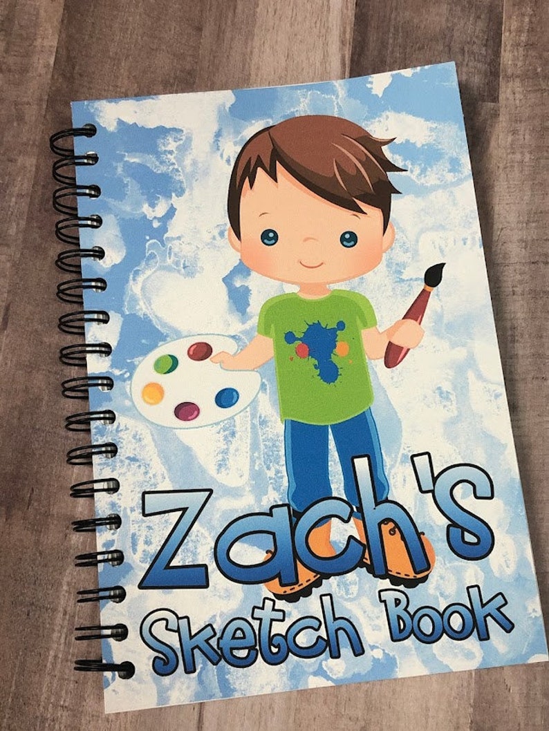 Op de afbeelding: Een spiraalgebonden schetsboek met een blauw en wit hemelthema. De omslag toont een cartoonillustratie van een jongen met een penseel en een verfpalet. De tekst "Zach's Sketch Book" staat op de omslag.