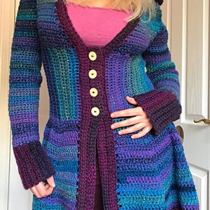 May include: A long, crocheted cardigan with a hood, featuring a patchwork design in shades of purple, blue, and green. The cardigan has a V-neckline and is fastened with buttons.