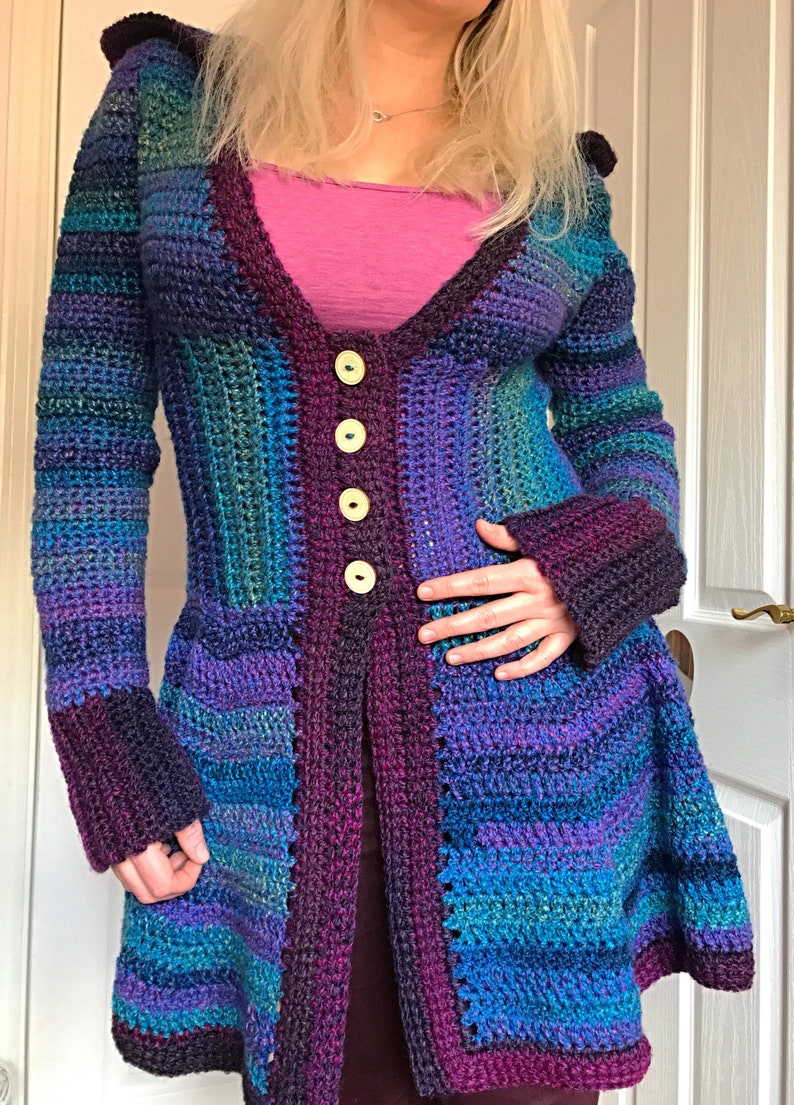 May include: A long, crocheted cardigan with a hood, featuring a patchwork design in shades of purple, blue, and green. The cardigan has a V-neckline and is fastened with buttons.