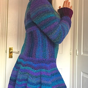 May include: A long, hooded crocheted cardigan with a striped pattern in shades of purple, blue, and teal. The cardigan has a flared hem and is made with a soft, textured yarn.