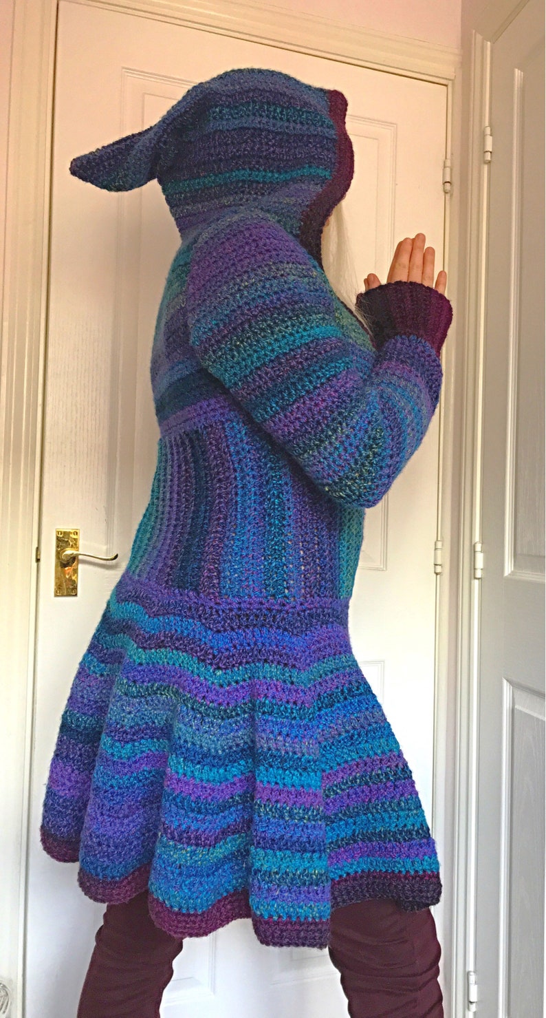 May include: A long, hooded crocheted cardigan with a striped pattern in shades of purple, blue, and teal. The cardigan has a flared hem and is made with a soft, textured yarn.