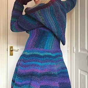 May include: A long-sleeved, hooded crocheted cardigan with a striped pattern in shades of purple, blue, and green. The cardigan has a flared hem and is worn over purple pants.