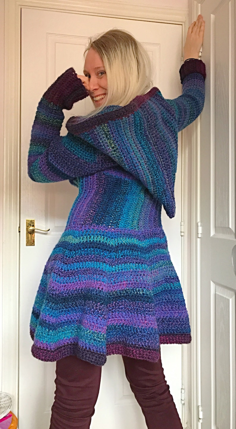 May include: A long-sleeved, hooded crocheted cardigan with a striped pattern in shades of purple, blue, and green. The cardigan has a flared hem and is worn over purple pants.