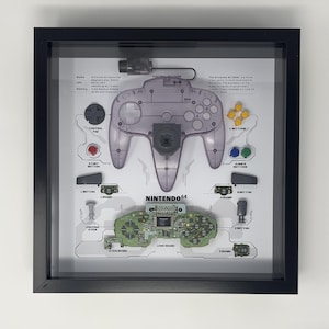 Puede incluir: Una vitrina enmarcada de un mando de Nintendo 64 desmontado. El mando es transparente y muestra los componentes internos. La pantalla también incluye la placa base y otras partes de la consola. El texto "NINTENDO 64" está impreso en la pantalla.