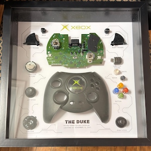 Może przedstawiać: Oprawiona prezentacja rozebranego oryginalnego kontrolera Xbox, "The Duke". Zawiera zieloną płytkę drukowaną, szarą obudowę kontrolera, przyciski, spusty i silniczki wibracyjne. Widoczny tekst "THE DUKE" i data premiery.