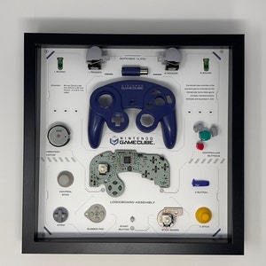 May include: Framed Nintendo GameCube controller parts display. Includes a blue controller, logic board, buttons, and other components. The display is in a black frame and features text labels for each part.