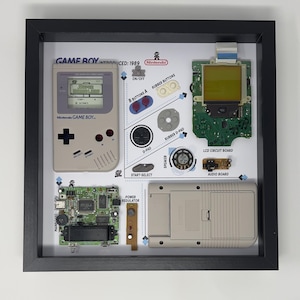 May include: A framed display of a disassembled Game Boy, showing the individual components of the handheld gaming console. The display includes the mainboard, LCD circuit board, audio board, power regulator, speaker, and buttons. The text "GAME BOY INTRODUCED: 1989" is visible at the top of the display.