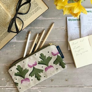 May include: A canvas pencil case with a radish design, open book, glasses, pencils, and daffodils on a wooden surface. The pencil case has a green zip and the text "LOVELY LYDIA". A handwritten note lists plants.