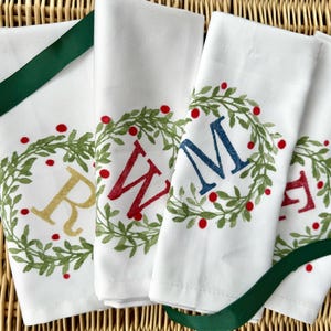 May include: White linen hand towels with embroidered wreaths and initial letters in gold, red, and blue. Each towel features a green wreath with red berries and a green ribbon. The towels are arranged on a woven wicker surface.