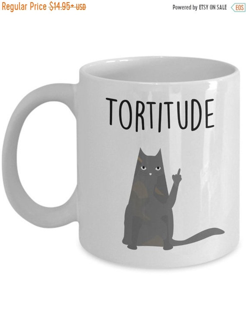 以下が含まれることがあります： 白いセラミック製のコーヒーカップに、黒い猫のイラストと「Tortitude」という文字が書かれています。猫は中指を立てて座っています。