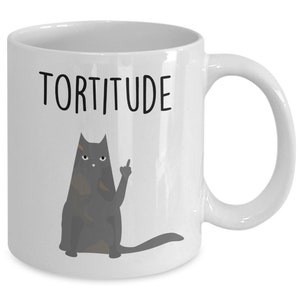 以下が含まれることがあります： 白いセラミック製のコーヒーカップ。黒い猫のイラストが中指を立てており、黒い文字で「Torttitude」と書かれている。